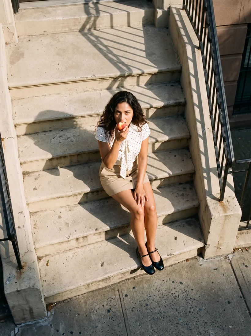 Model in a polka-dot tie-neck blouse and khaki button mini skirt and Mary Janes seated on a stone stoop, seen from above in hard sun, generated with Gemini Pro and a Uwear art direction