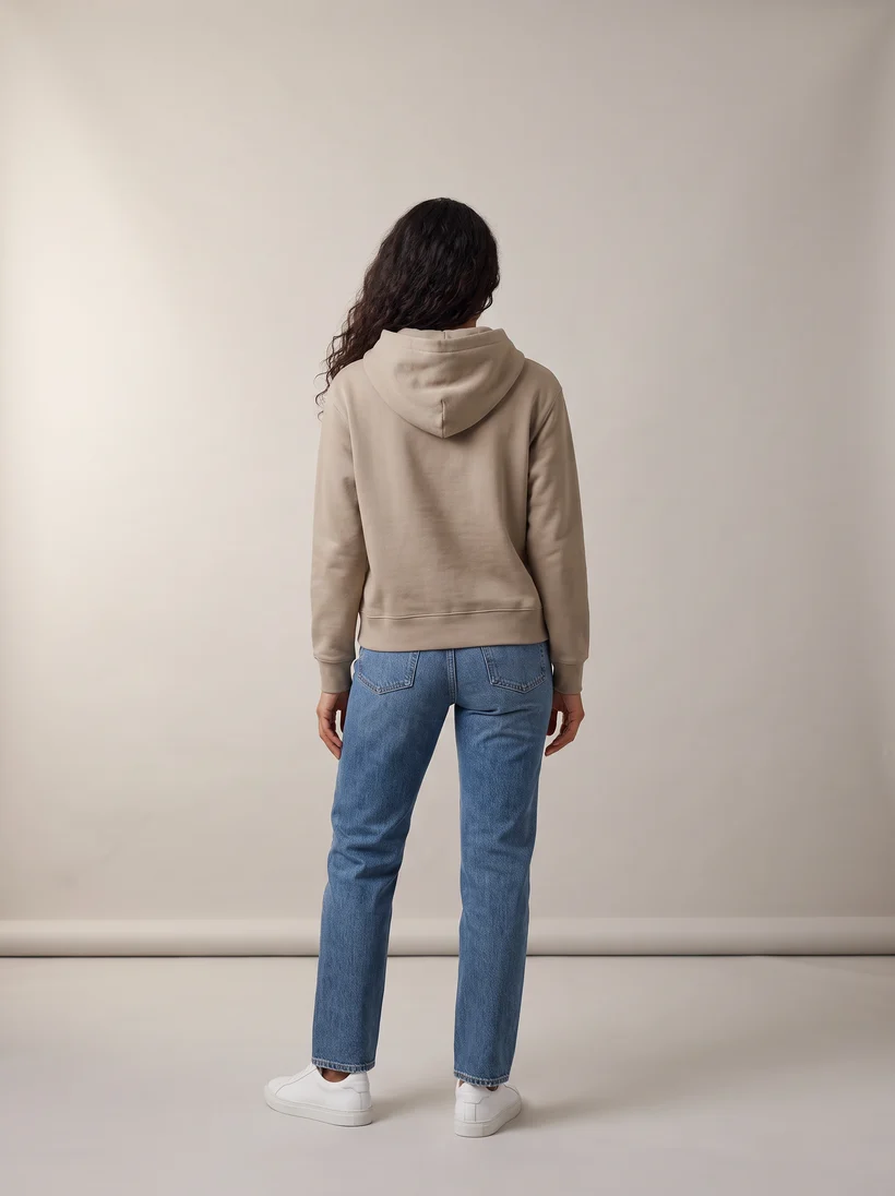 The same model seen from behind in the taupe hoodie and jeans on the grey paper backdrop, the back pose of the original shoot, generated with Gemini Pro