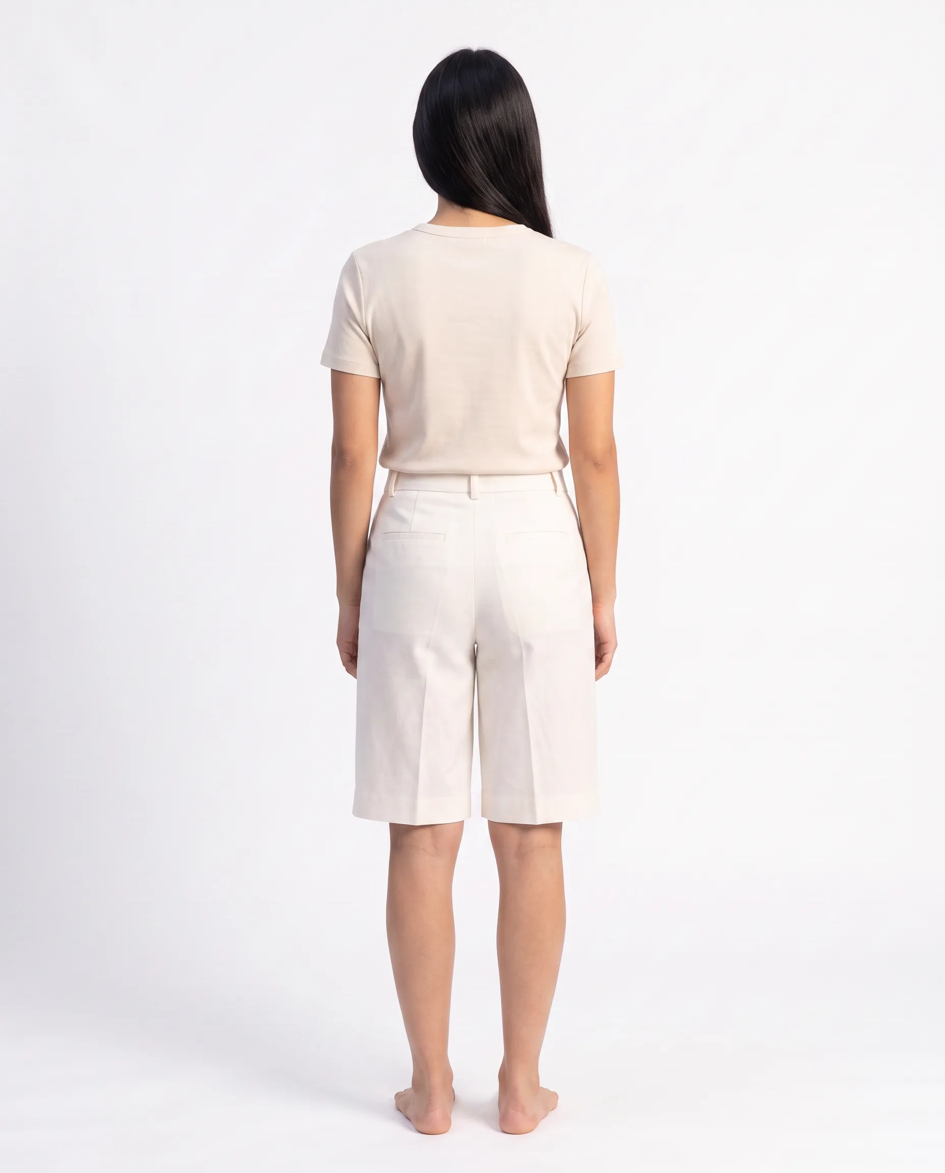 Back view of a female model wearing the same cream tailored knee-length shorts with a relaxed drapey fit against a studio backdrop