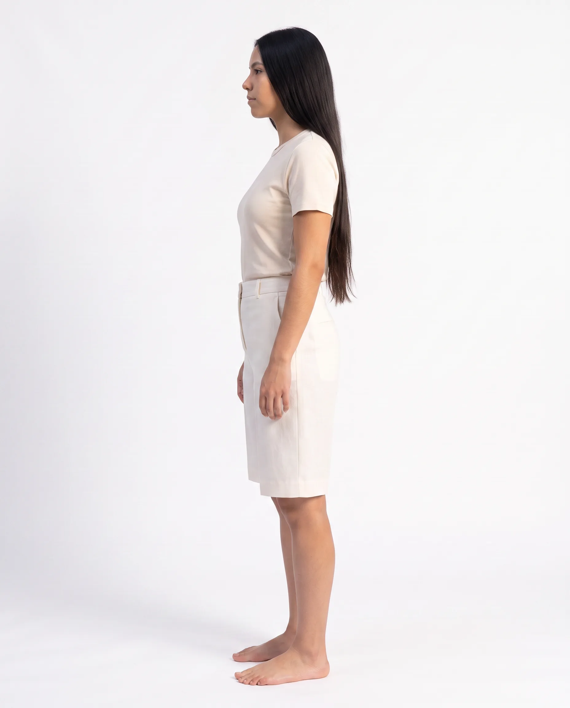 Side view of a female model wearing the same cream tailored knee-length shorts with a relaxed drapey fit against a studio backdrop